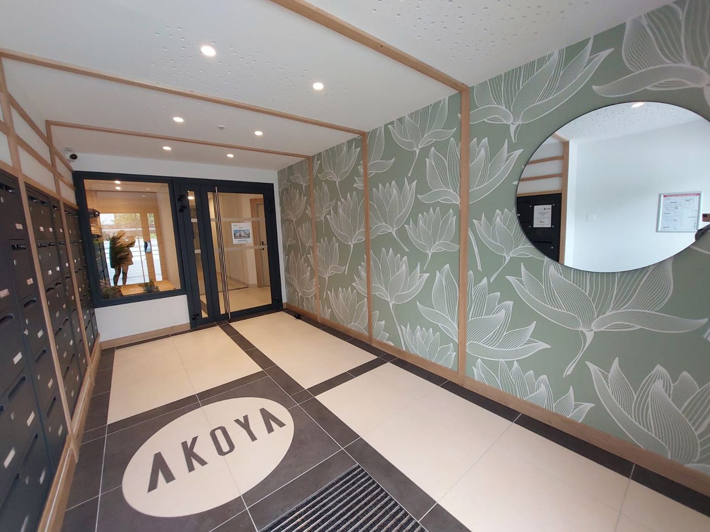 Akoya à Bayonne : hall d’entrée, papier peint végétal, miroir rond et logo au sol.