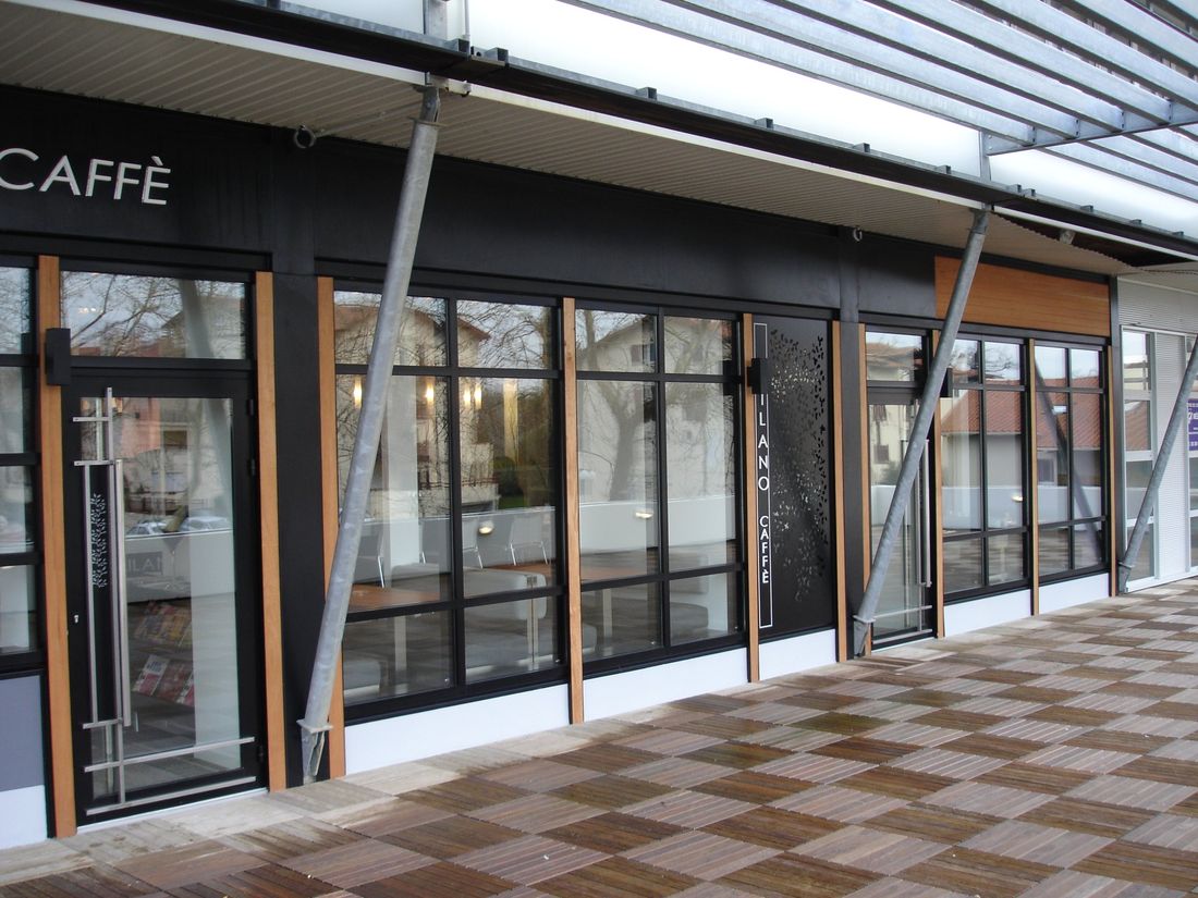 Façade d’un café-restaurant : menuiseries noires, montants bois, panneau métallique découpé, terrasse en caillebotis.