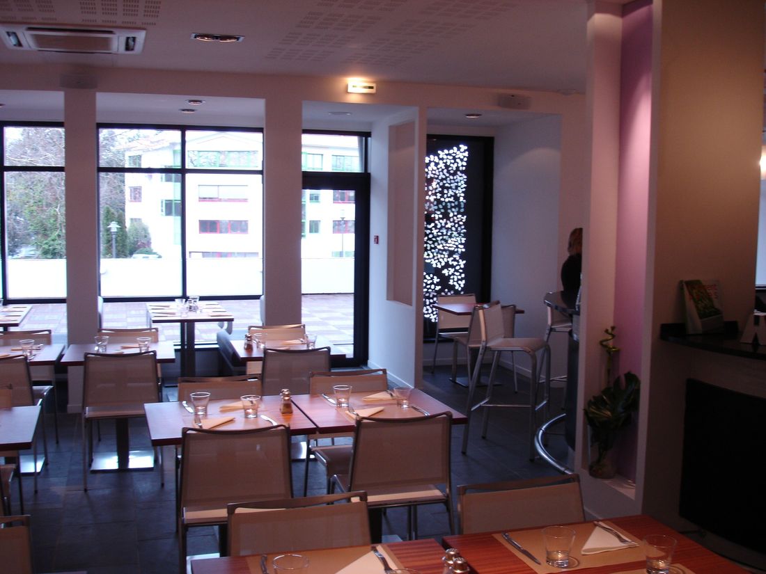 Salle d’un café-restaurant : tables en bois, grandes baies vitrées sur la terrasse, panneau lumineux découpé.